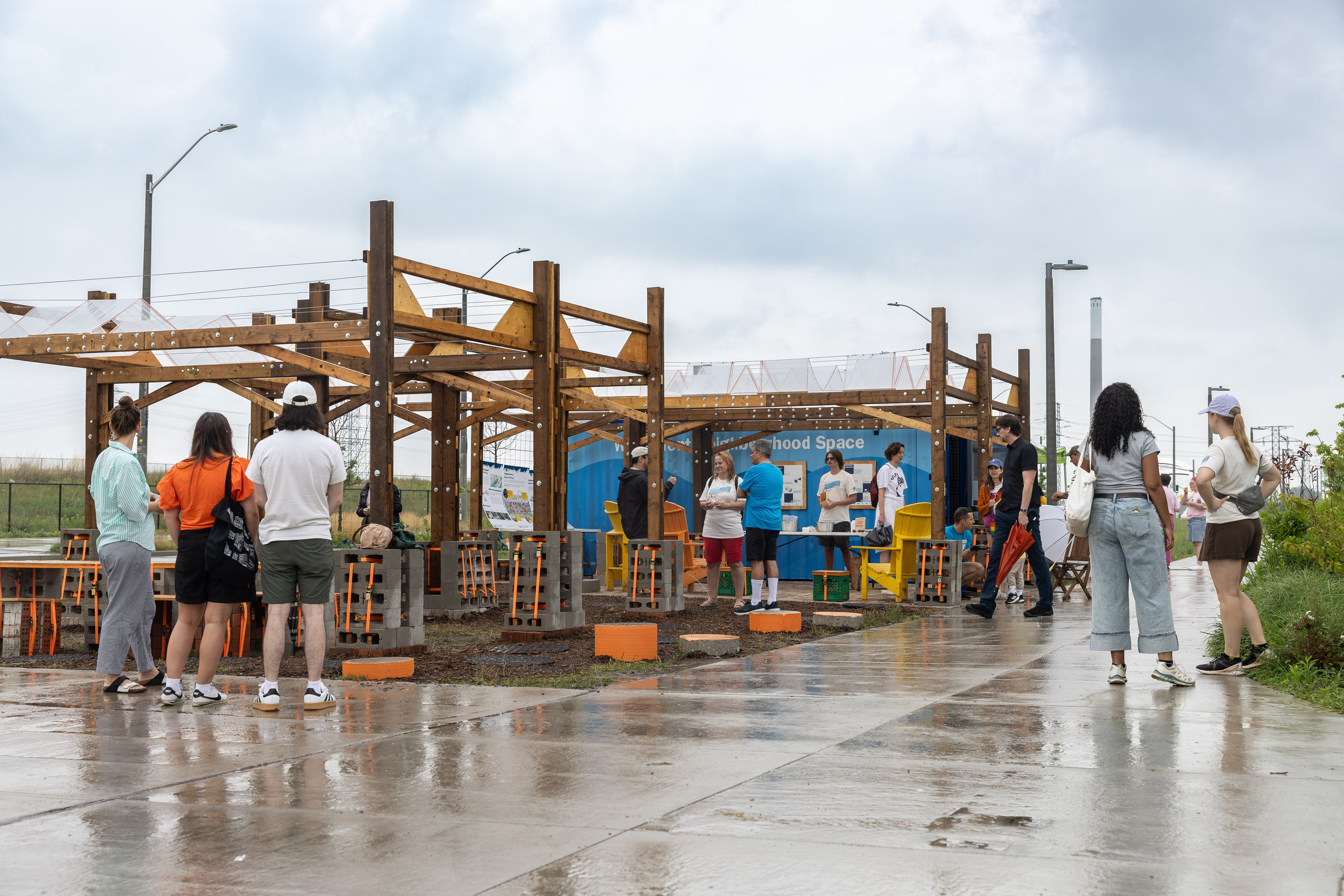 Visitors gather at an outdoor community space on a rainy day. Several people stand and walk along a wet concrete pathway beside a partially constructed wooden pavilion. A blue shipping container with informational displays serves as a backdrop, while chairs, tables, and construction materials are arranged throughout the site. Overcast skies and reflective pavement emphasize the rainy weather.