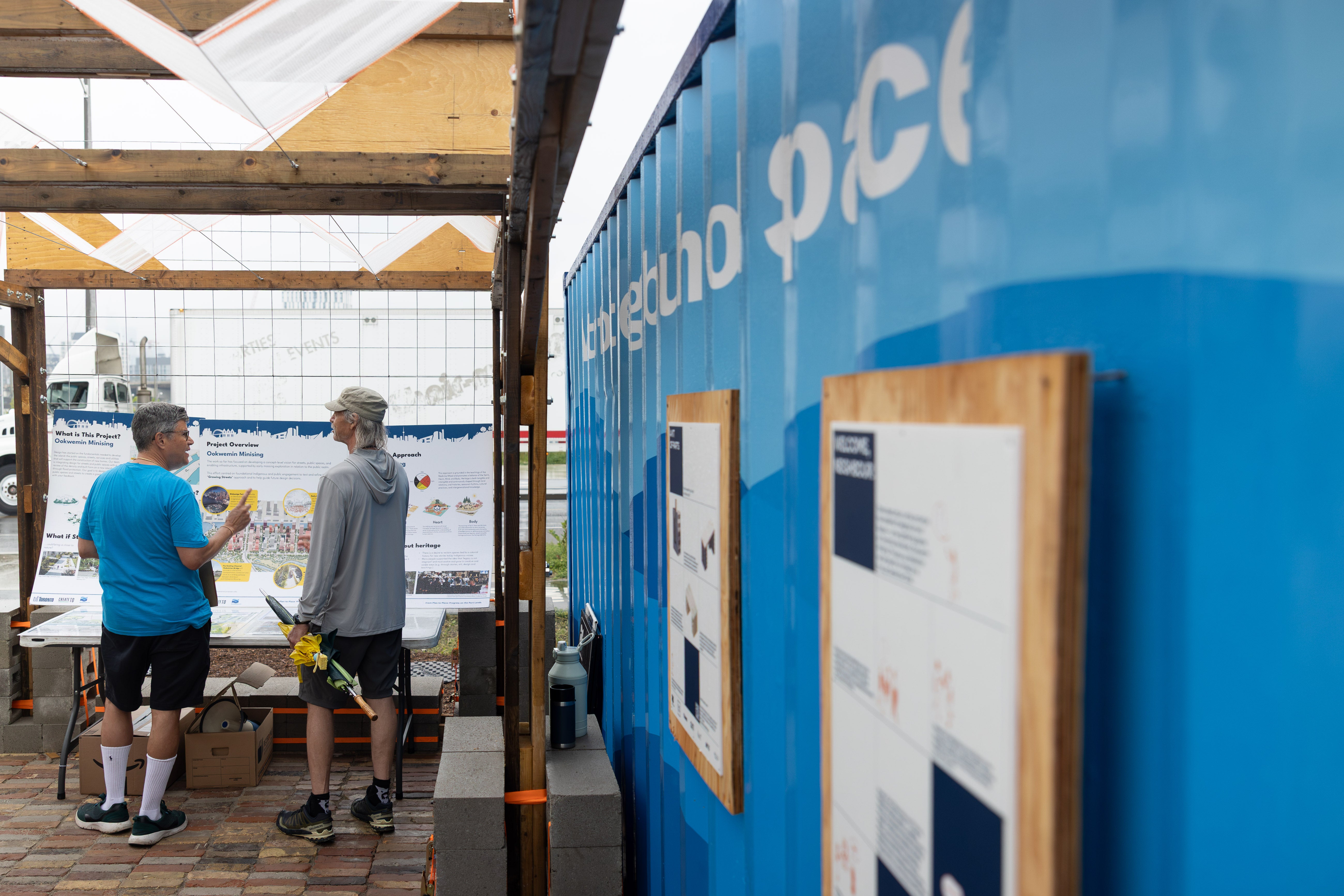 Two visitors stand inside a covered outdoor community space, reviewing project information boards mounted beneath a timber pavilion. A large blue shipping container displaying the words “Neighbourhood Space” forms one side of the structure, with additional educational panels attached to its exterior. The ground is wet from recent rain, and information displays about the Ookwemin Minising project are visible in the background.