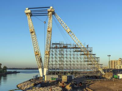 A massive crane with scaffolding stands tall on the edge of river in the middle of a construction site. 