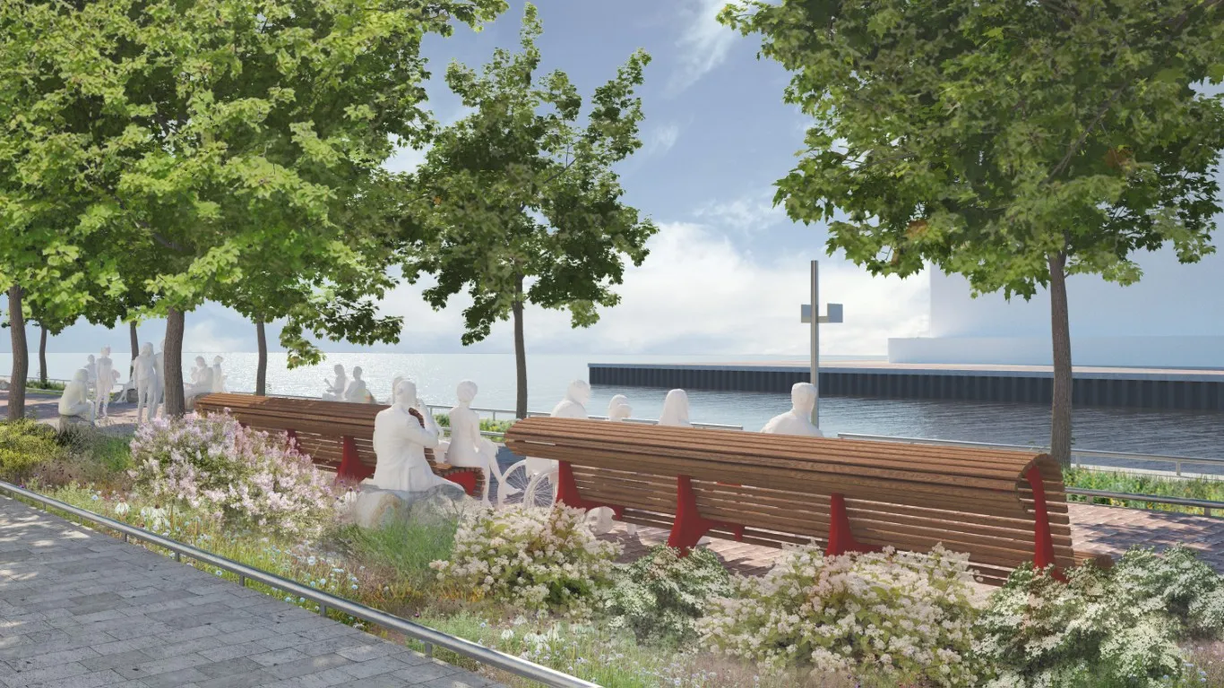 A row of long wooden benches faces Lake Ontario along the Water's Edge Promenade, shaded by trees and surrounded by lush planting. White sculpted figures, including one in a wheelchair, occupy the benches and adjacent companion seating areas.