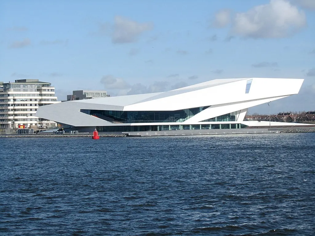 An angular white building—the EYE Filmmuseum in Amsterdam—sits prominently at the edge of a waterfront, with expansive glass windows and modern geometric design. Surrounded by water and adjacent residential buildings, the museum serves as a precedent for how a cultural destination can activate the water's edge, illustrating potential for a similar cluster of cultural and community uses at Toronto's waterfront.
