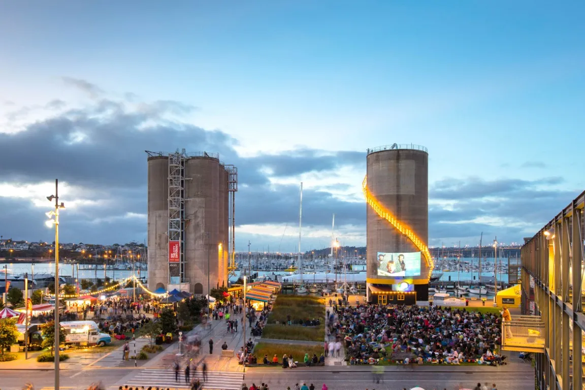 A lively waterfront space at Silo Park in Auckland, New Zealand, where a crowd watches a movie projected onto a silo. Decorative string lights spiral up the silo, and people gather around food stalls and a grassy lawn. This precedent image illustrates how a silo-adjacent park can support cultural programming and community use.