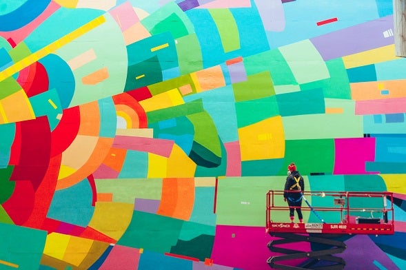 an artist on a scaffold painting a bright and colourful mural on a building