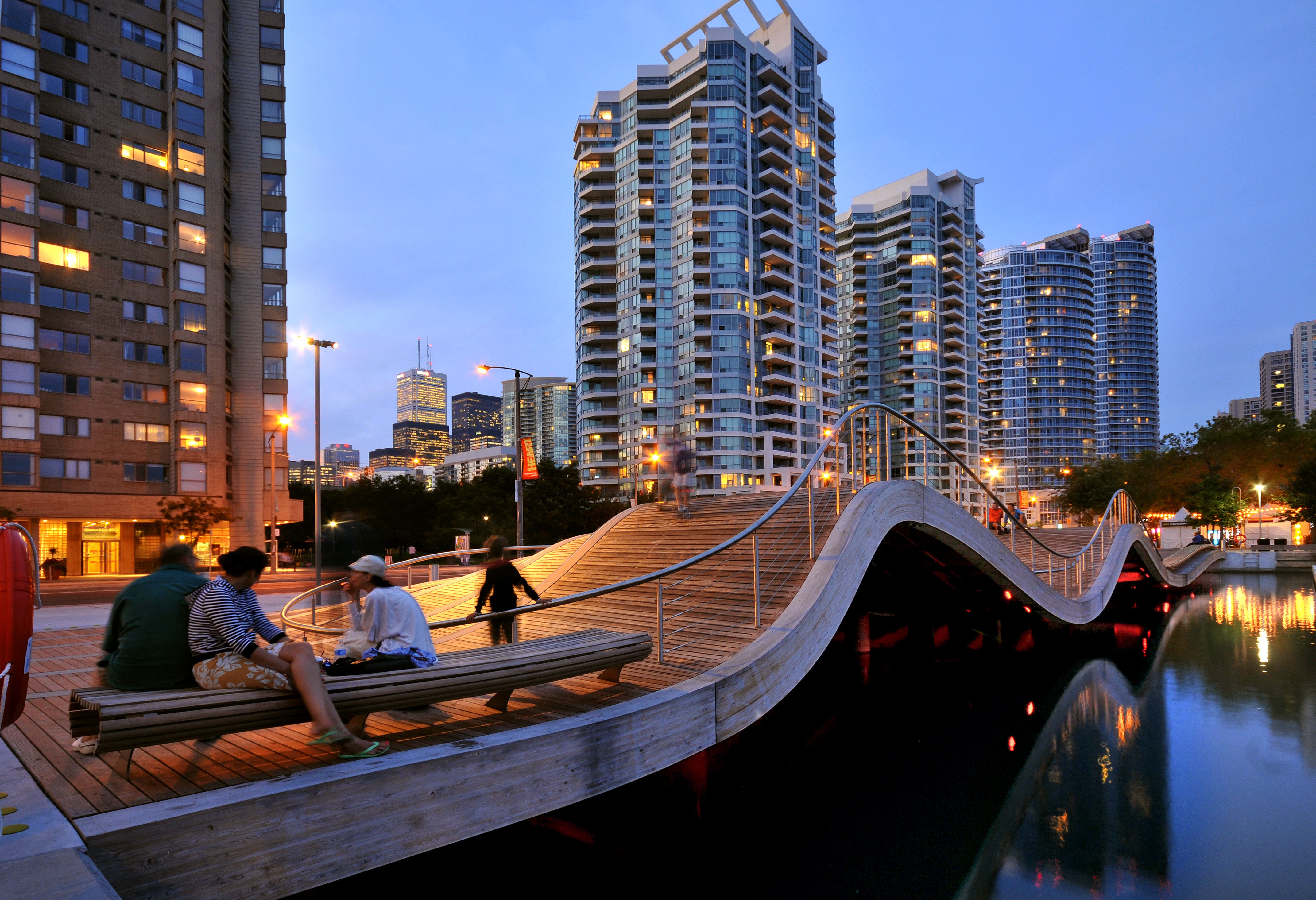 people sitting along the wavedecks benches next to Lake Ontario at dusk - buildings in the background