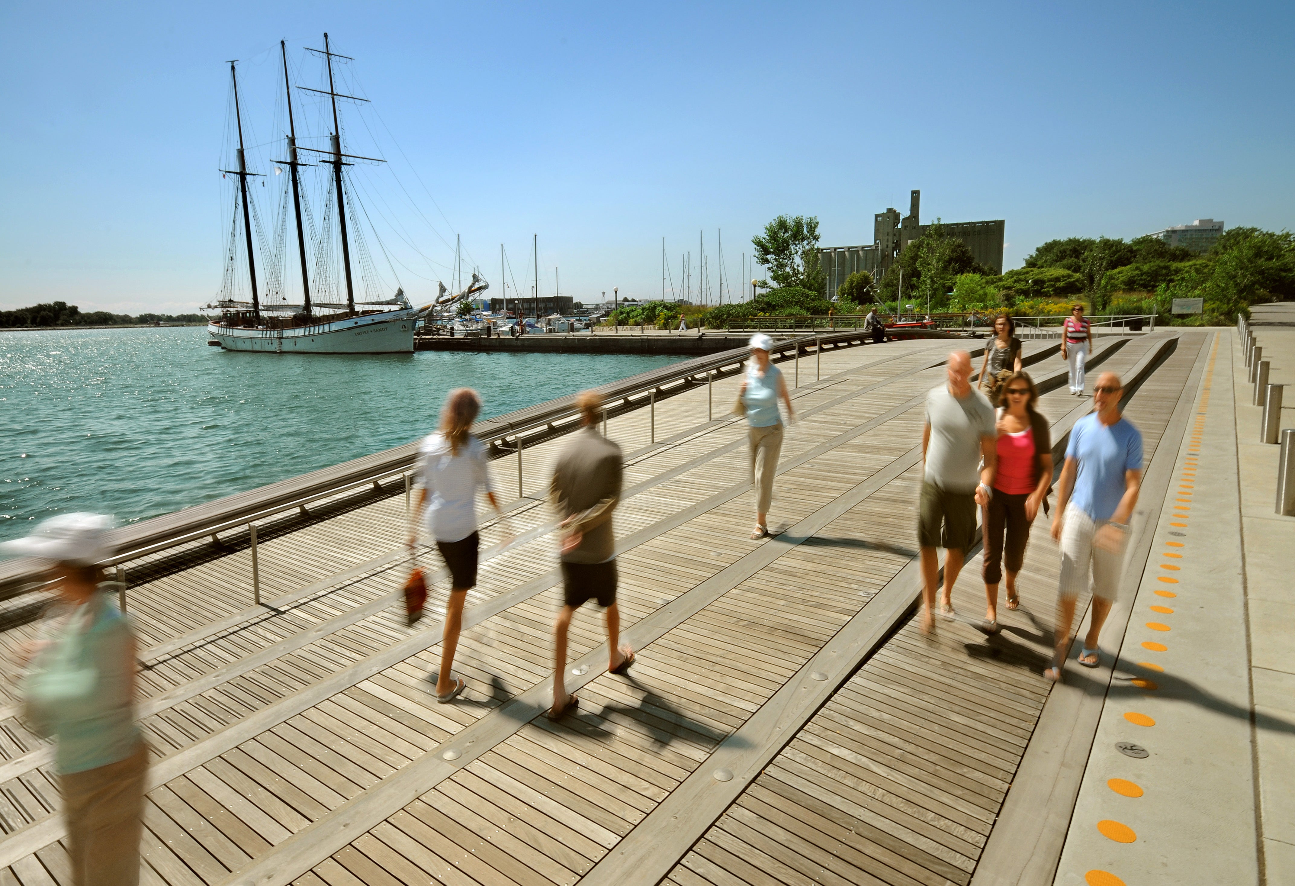 people walking along the Spadina WaveDeck next to Lake Ontario with a sail boat in the background