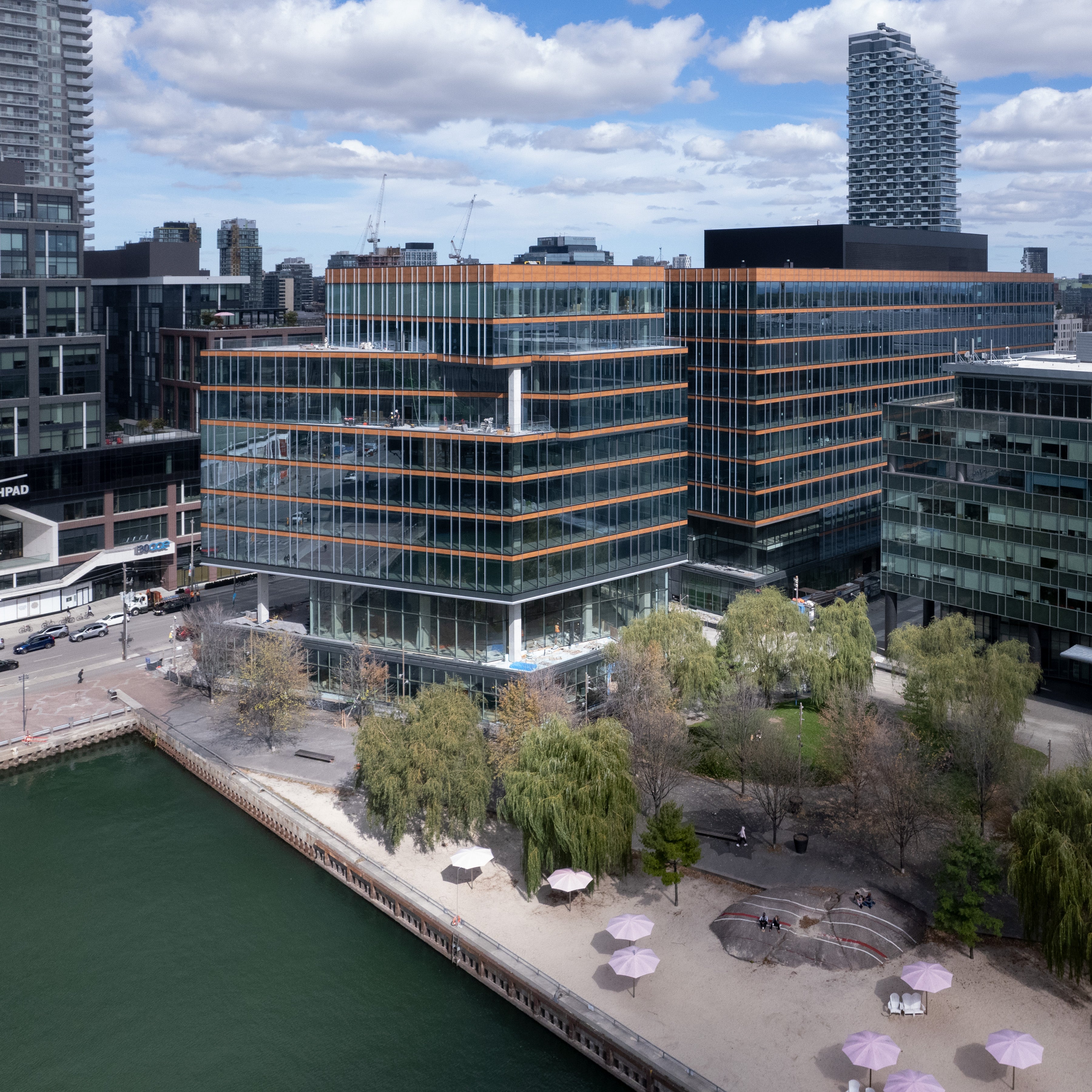 An angular blue glass building next to a park with weeping willow trees, pink umbrellas and a white sand beach.