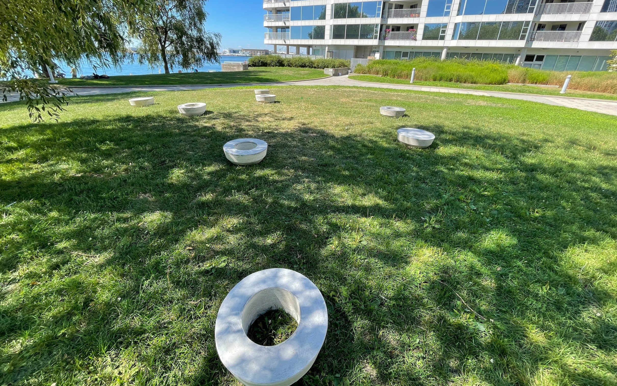 A sculpture with eight separate concrete rings on a grassy lawn. An apartment building and lake in the background.