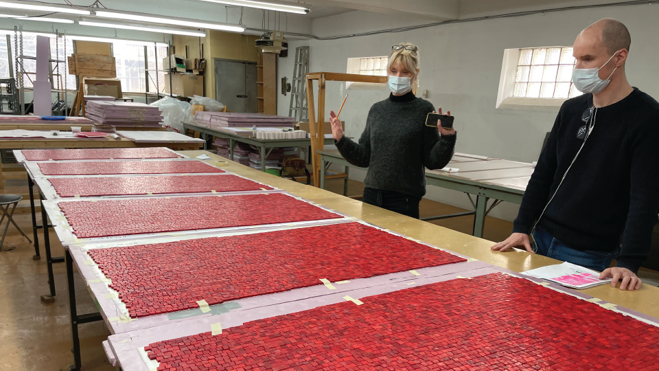 In a workshop, two stand behind a long table with red tiles.