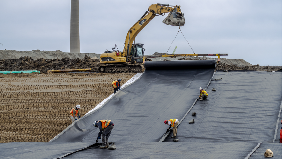 Construction workers lay out a strip of black plastic on a dirt ground. Excavator in the background. 