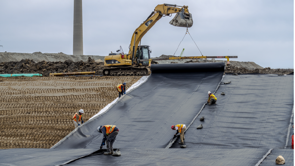 Construction workers lay out a strip of black plastic on a dirt ground. Excavator in the background.