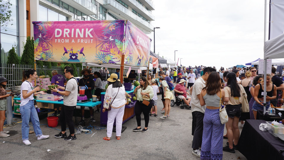 A crowded outdoor food market, with a vendor sign reading 'Drink from a fruit'