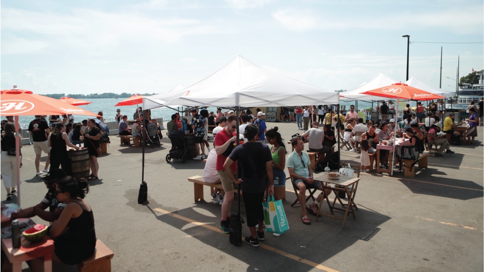 Groups of people eating food under umbrellas at an outdoor food market on a sunny day.