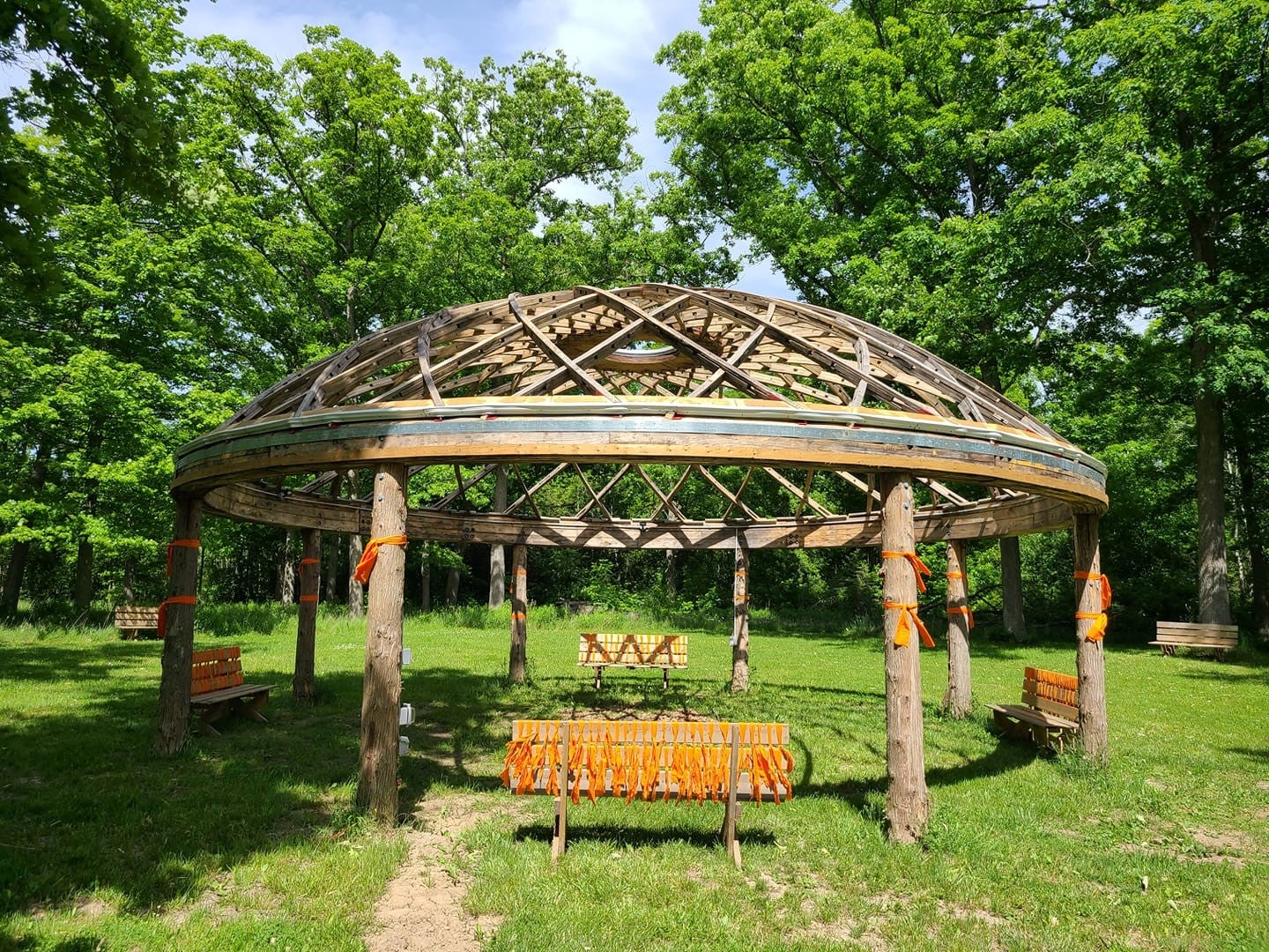 A wooden gathering space. The circular structure has four benches and orange decorative ties. 