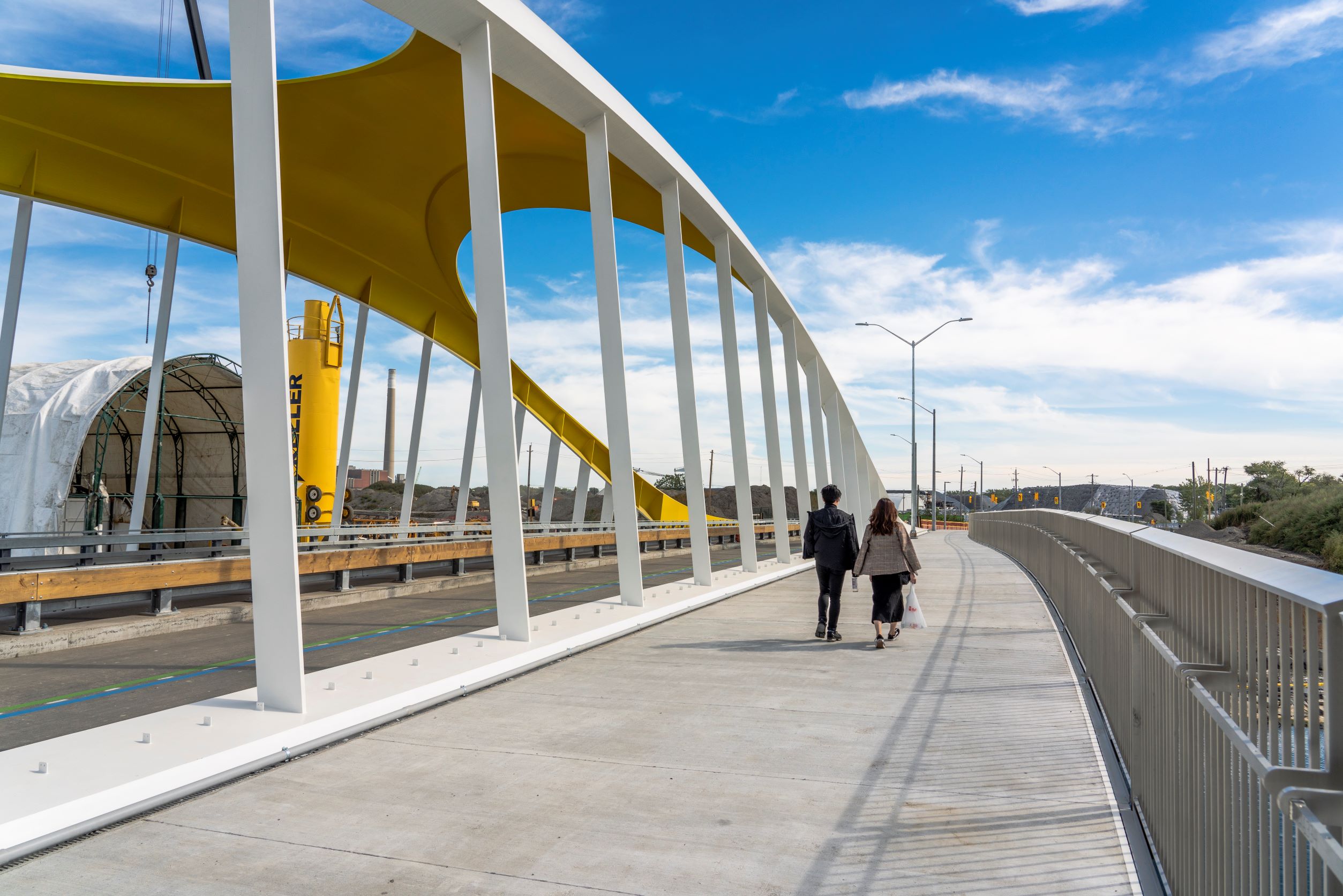 Two people walk along a wide sidewalk next to the yellow and white arch of the bridge.