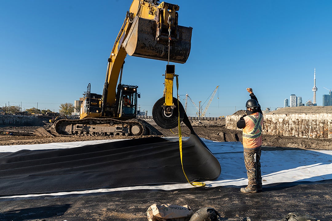 An excavator is holding up a large roll of black material, which is being unrolled to cover the ground.