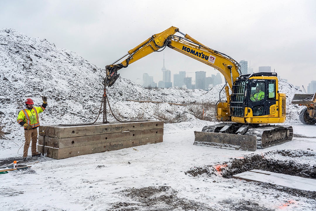 An excavator lifts large rows of plywood on a snowy day.