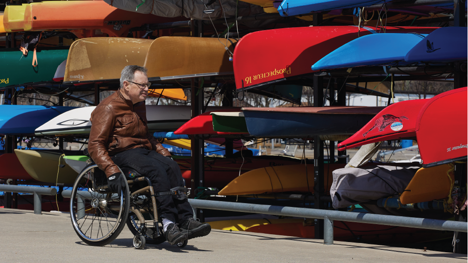 A person in a wheelchair, with canoes and kayaks in the background. 