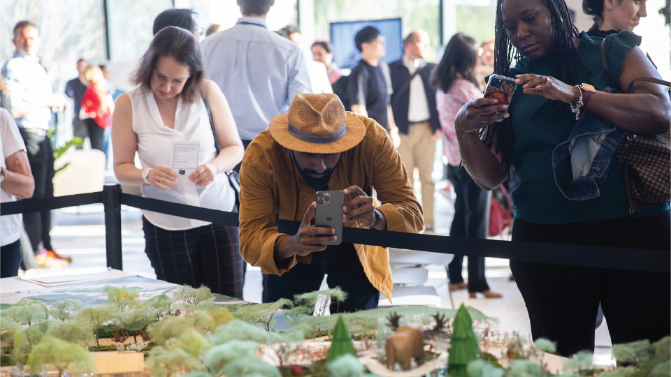 Three people standing in front of a model. Two people take pictures with their phones.