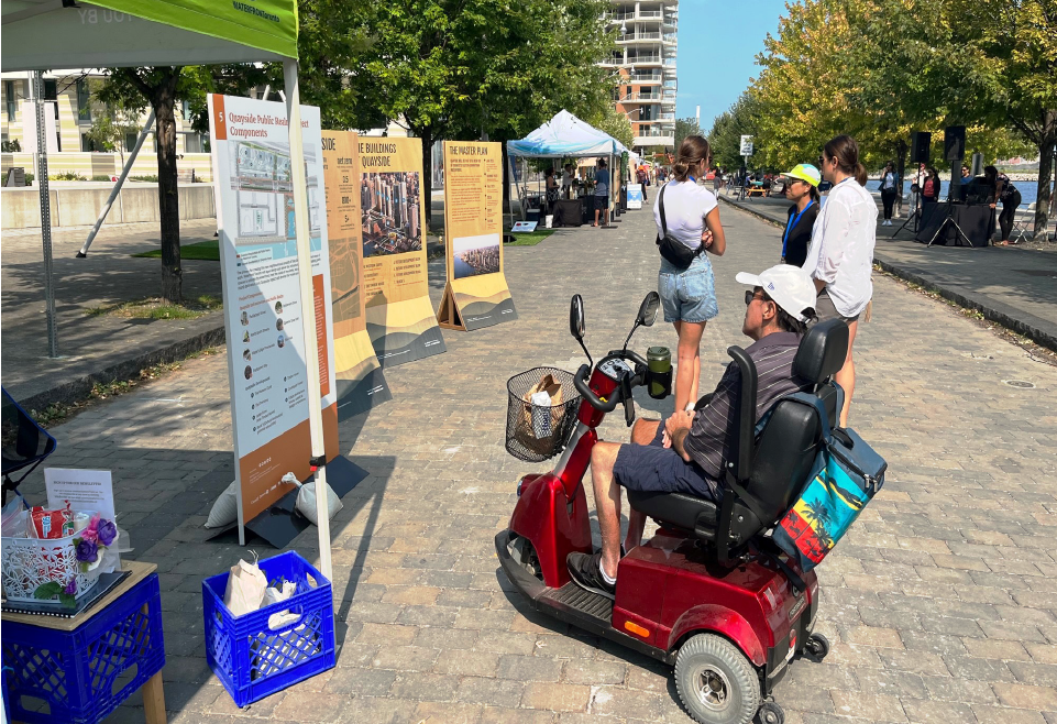 A person in a mobility chair reading a display board at a public event. 