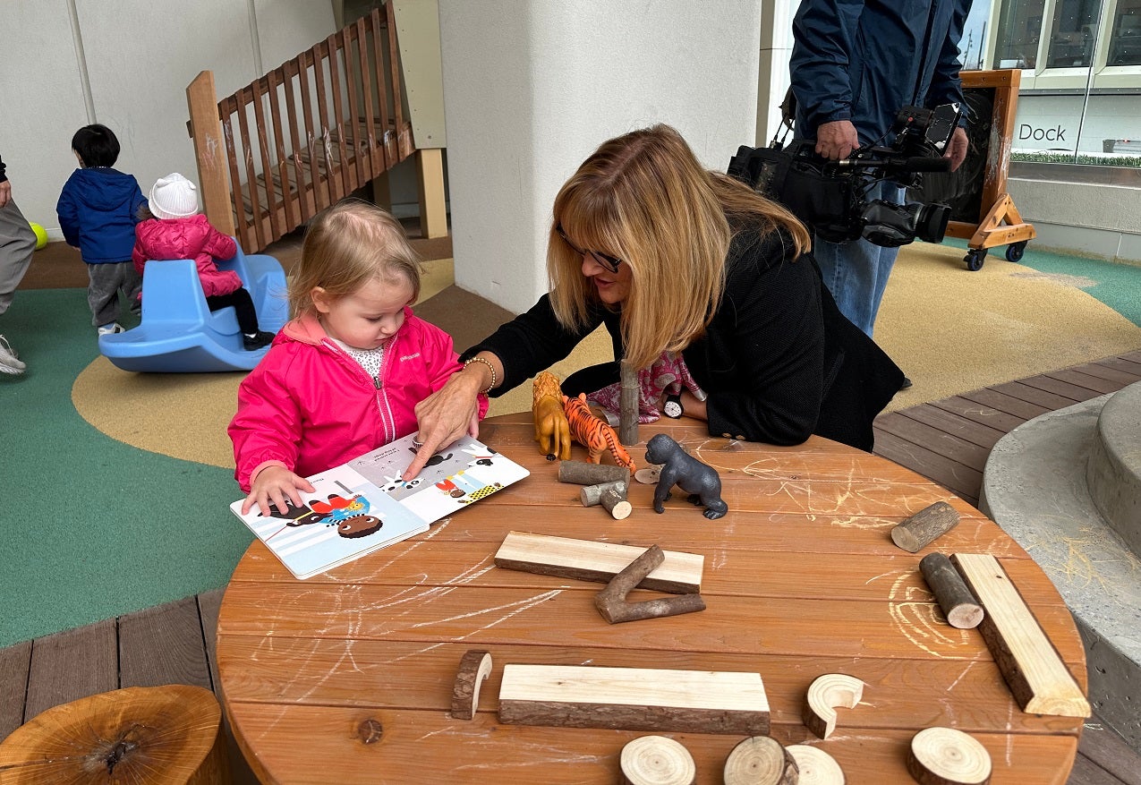 a woman sits at a small round table and reads to a toddler 