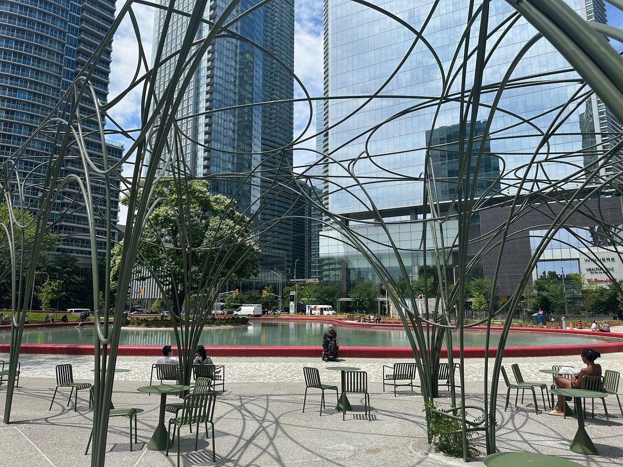 people sitting under a park's trellis and next to a large pond