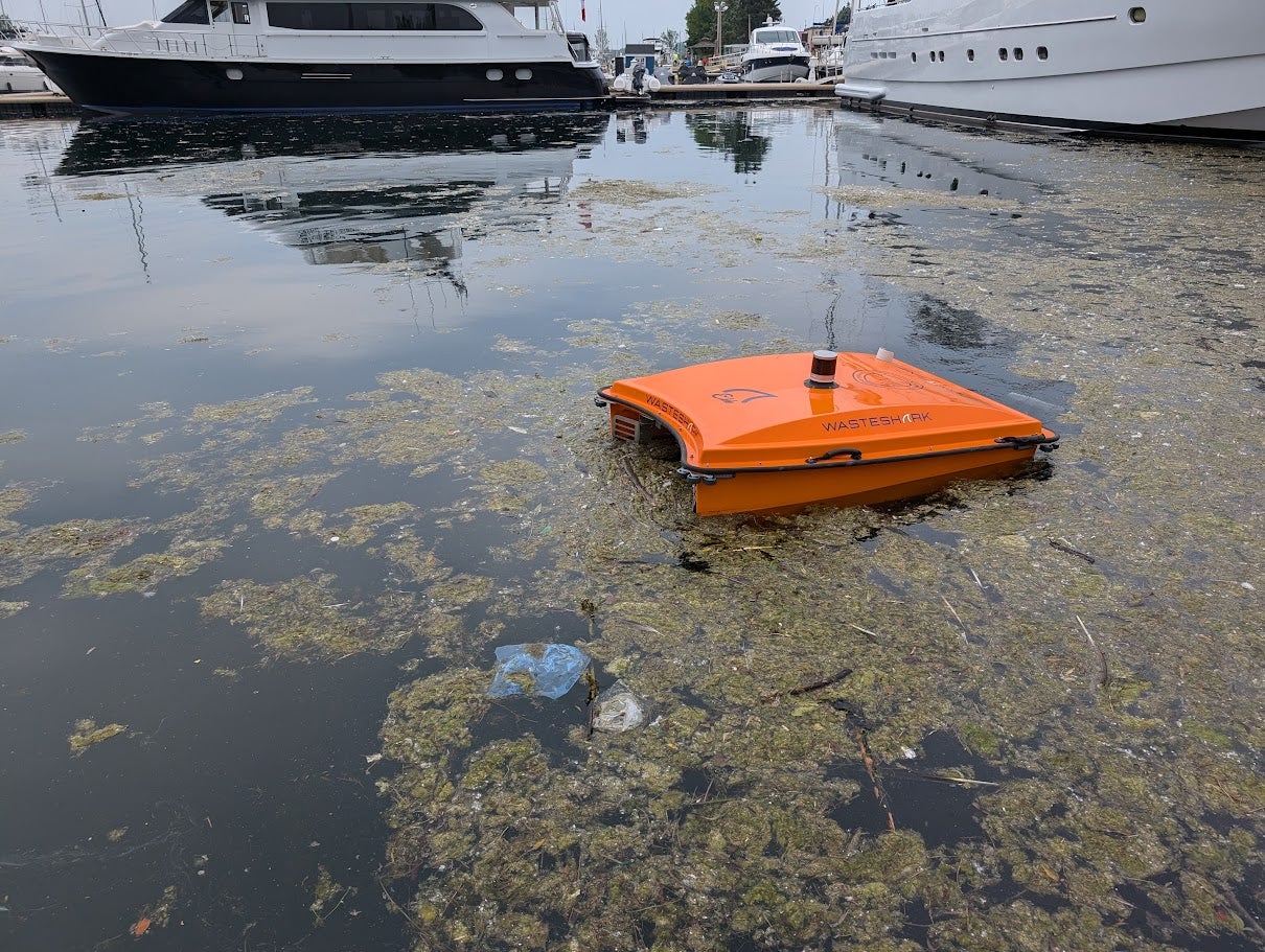 An orange WasteShark aquatic drone floating in a marina, surrounded by algae and debris, with large white yachts docked in the background.