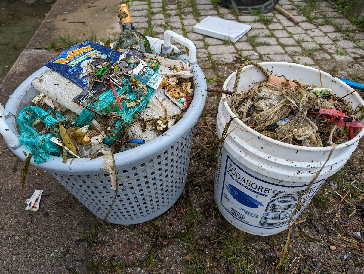Two buckets filled with assorted garbage including plastic wrappers, seaweed, and other debris, collected during a waterfront clean-up.