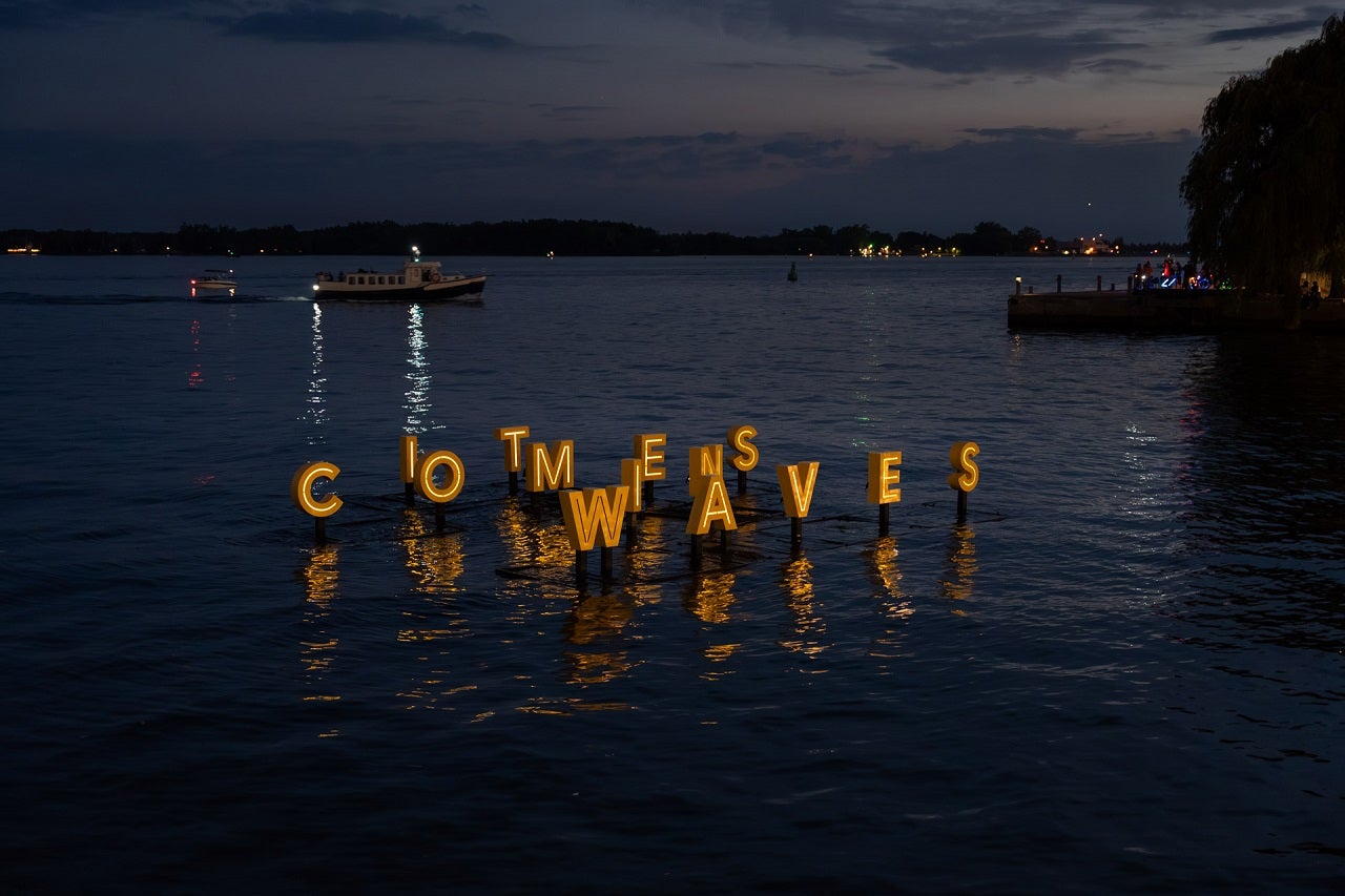 Floating artwork at night on water with big letters reads It Comes In Waves.