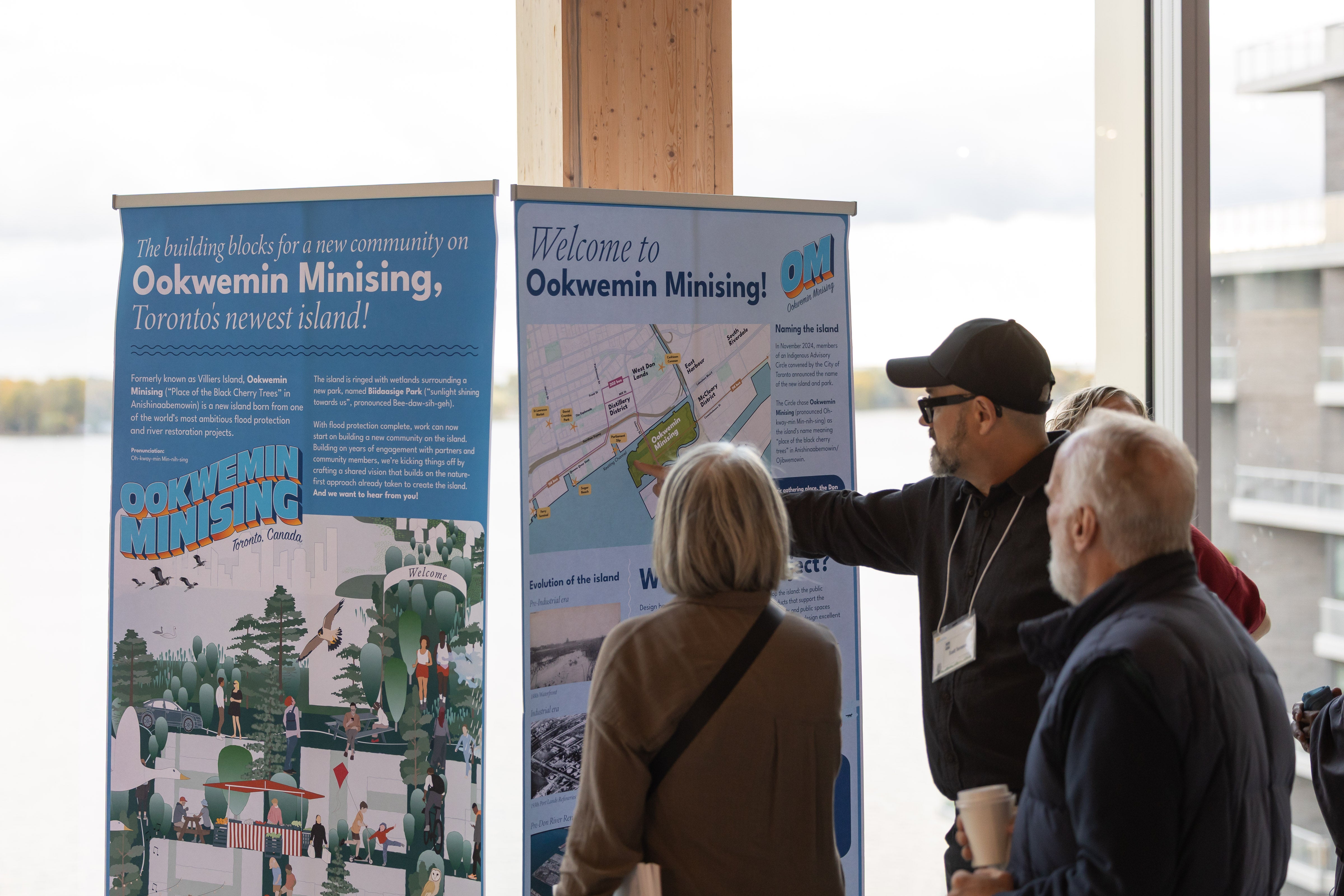 Several people stand indoors viewing two large informational display boards about Ookwemin Minising. 