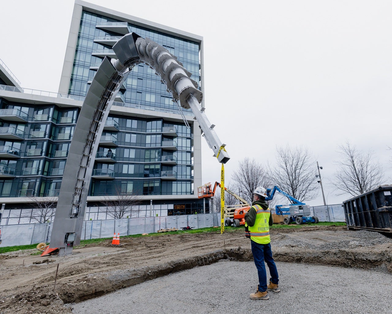 A crew member works on building a large "arch" public art piece behind fencing.