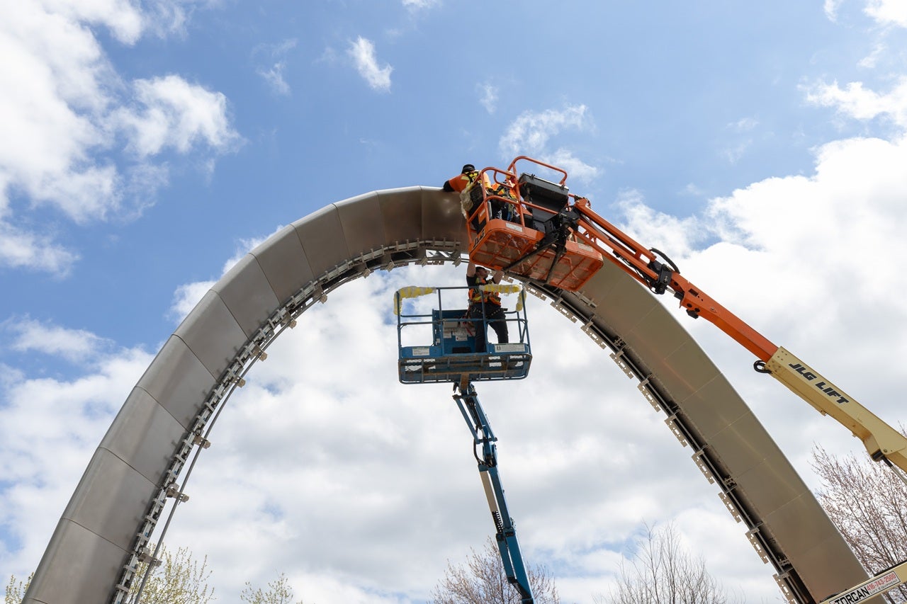 Crews use a large boom to reach the top of a large public art installation in the form of an arch.