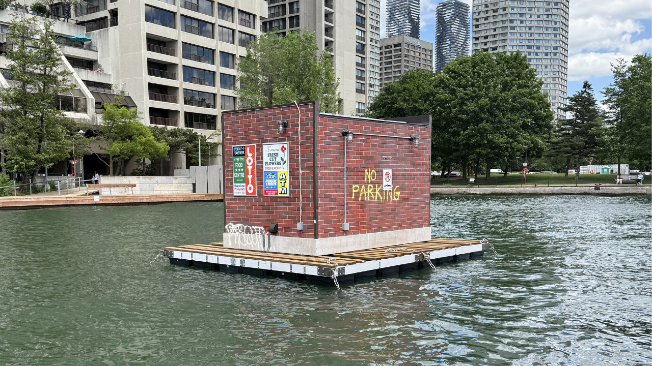 A small brick convenience store structure sits on a floating platform in the water, surrounded by buoy markers, with high-rise residential buildings and trees visible along the shoreline.