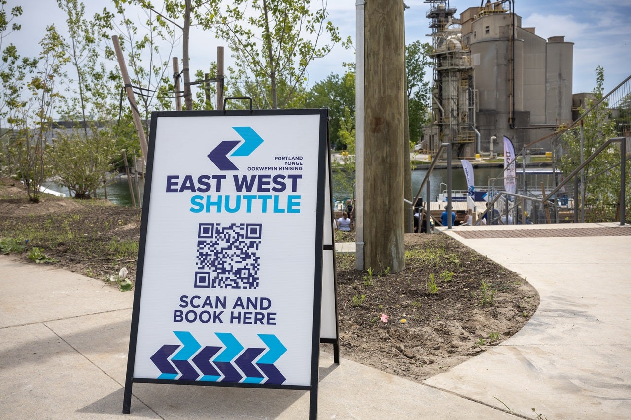 A sign board on a street that reads: East West Shuttle, scan and book here.