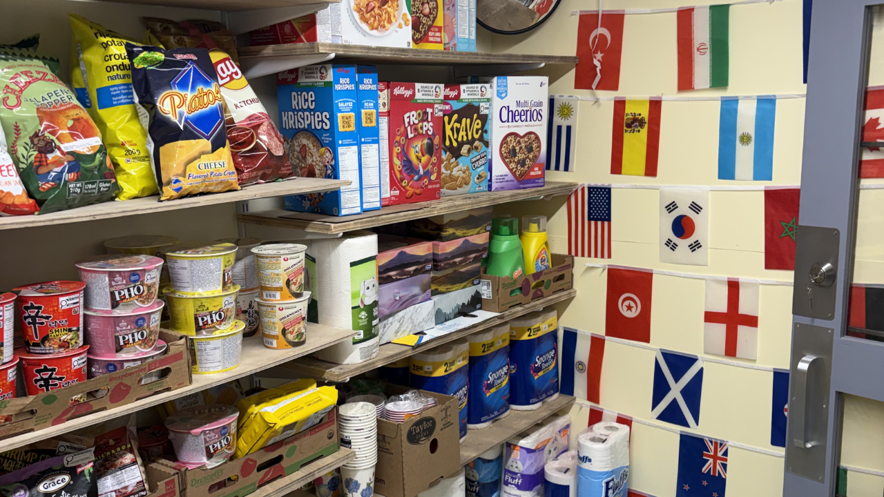 Interior shelves stocked with snacks, instant noodles, cereal, and household supplies, with a wall decorated in small international flags behind the display.