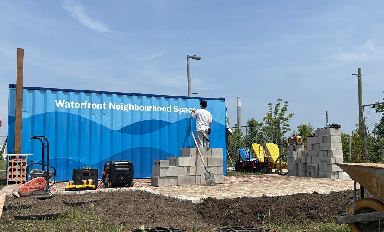 A person stencils on the words "Waterfront Neighbourhood Space" on to the side of a blue painted shipping container