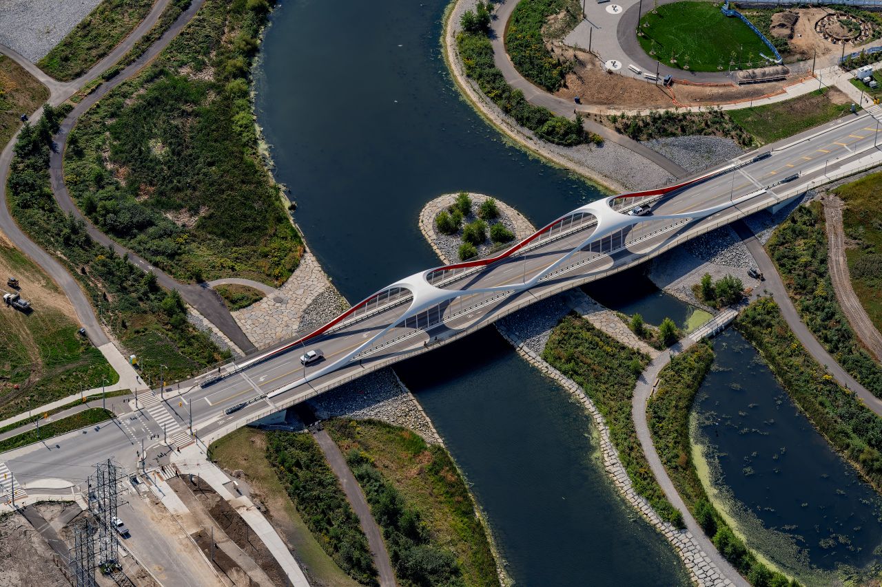 Aerial view of the Port Land Birdges at Cherry Street. The bridge features a modern design with interconnected oval shapes. Surrounding the river are green areas and pathways. Roads and vegetation line the riverbanks.