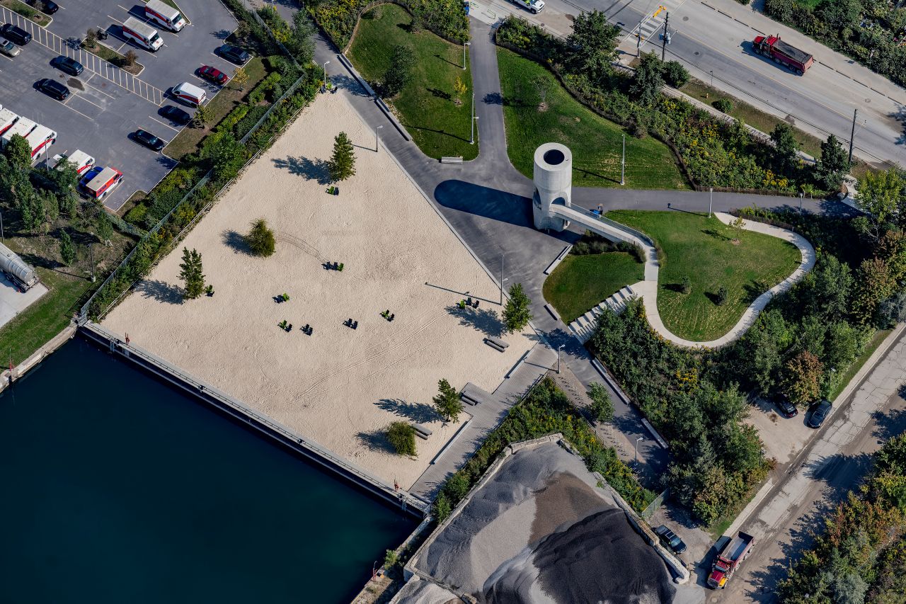 Aerial view of Leslie Lookout Park in Toronto, showcasing a sandy area with. To the right, a winding path leads past a circular, modern pavilion amidst landscaped greenery. Nearby roads and parked vehicles are visible, along with an adjacent industrial area.