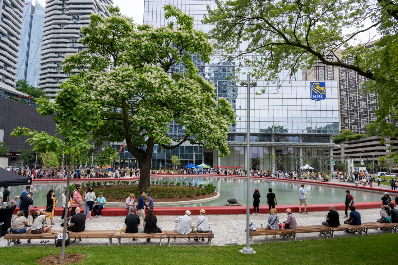 A busy outdoor park features a tree surrounded by benches where people are sitting and standing. In the background, a heart shaped pond is lined with more people. Tall buildings surround the plaza, one with the RBC logo visible.
