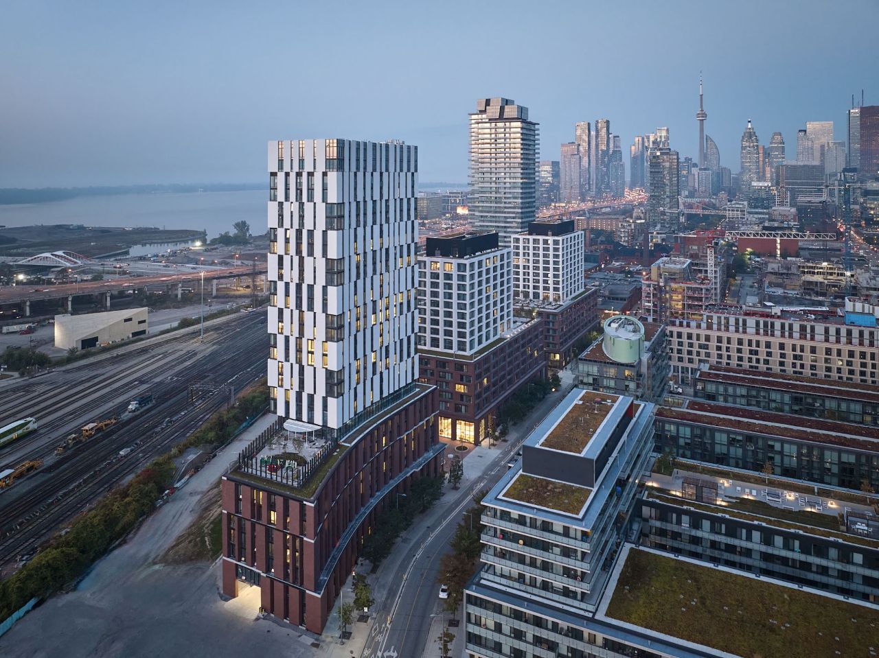 Aerial view of a modern urban area in Toronto during twilight. The landscape includes a distinctive high-rise building with a patterned facade, surrounded by lower structures with green rooftops. In the background, the city skyline features the CN Tower.