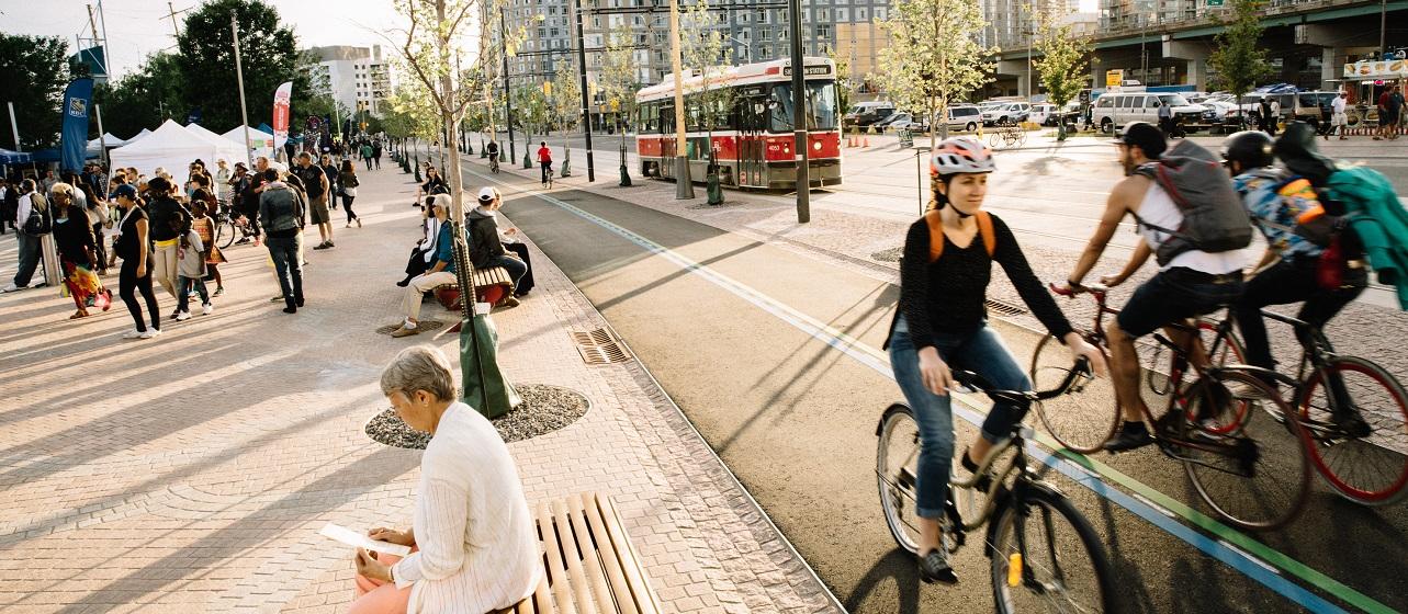 A bustling urban scene with people walking, sitting, and cycling next to streetcar tracks. A streetcard is in the background, and a cyclist in a helmet rides in the foreground. Trees and modern buildings line the street.