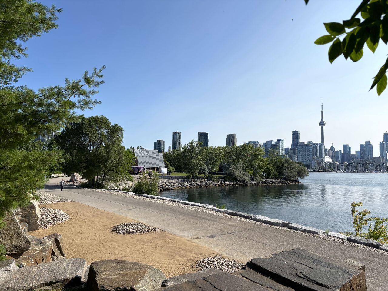 Toronto's skyline with CN tower, view of lake ontario on the right and greenery, trail, stones on the left.