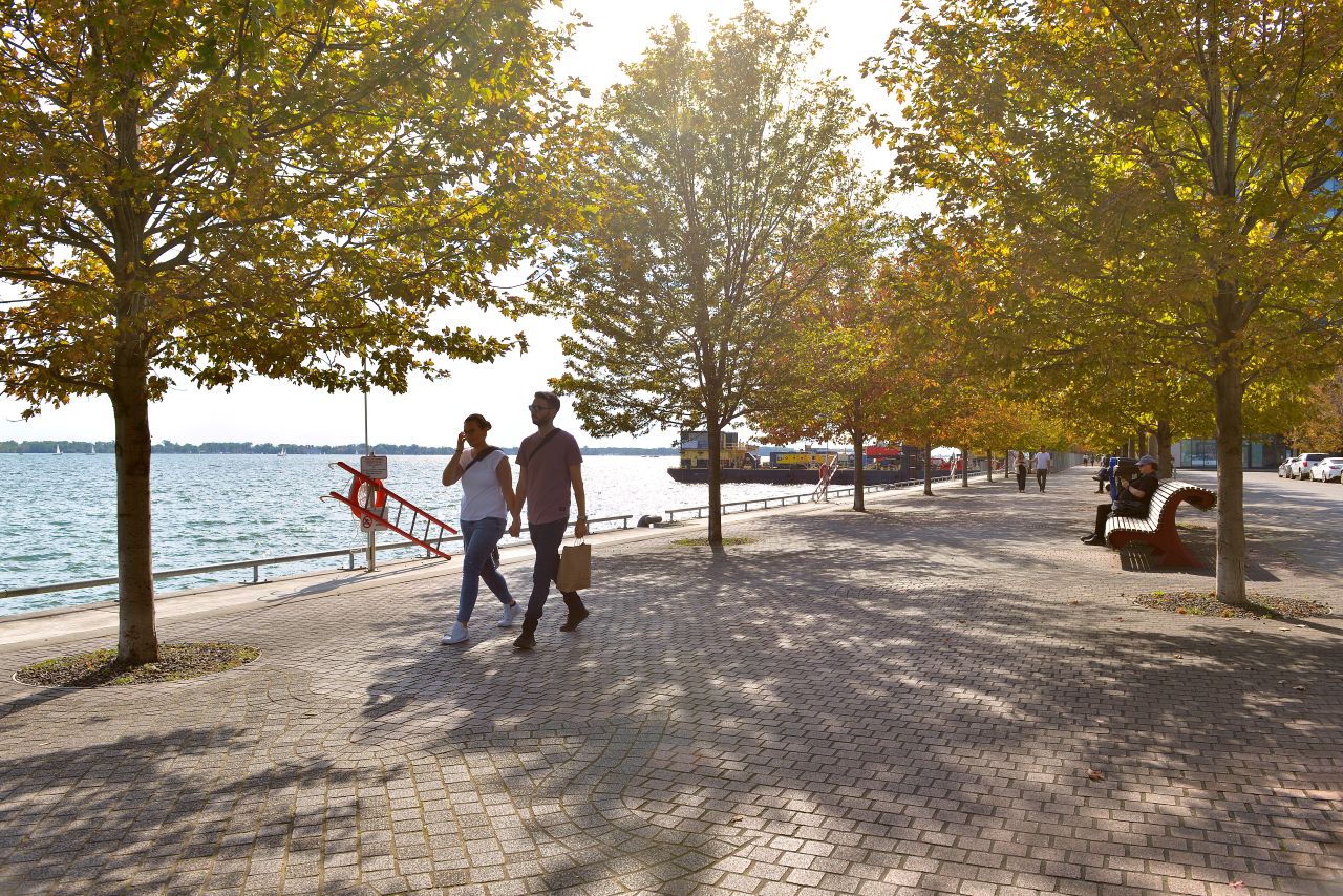 A scenic riverside walkway lined with trees on a sunny day. Two individuals stroll along the path, one carrying a shopping bag. A bench and a few parked bicycles are visible. In the background, a riverboat is docked on the water.