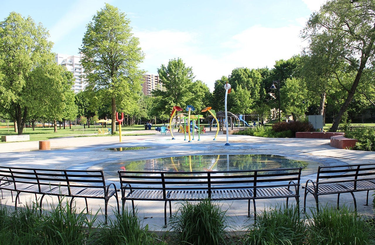Splash pad surfaces and multi-coloured play fixtures dot the Marie Curtis Park playground, which is surrounded by greenery and outlined with seating areas. (Image by Pkvan, from Wikimedia Commons)