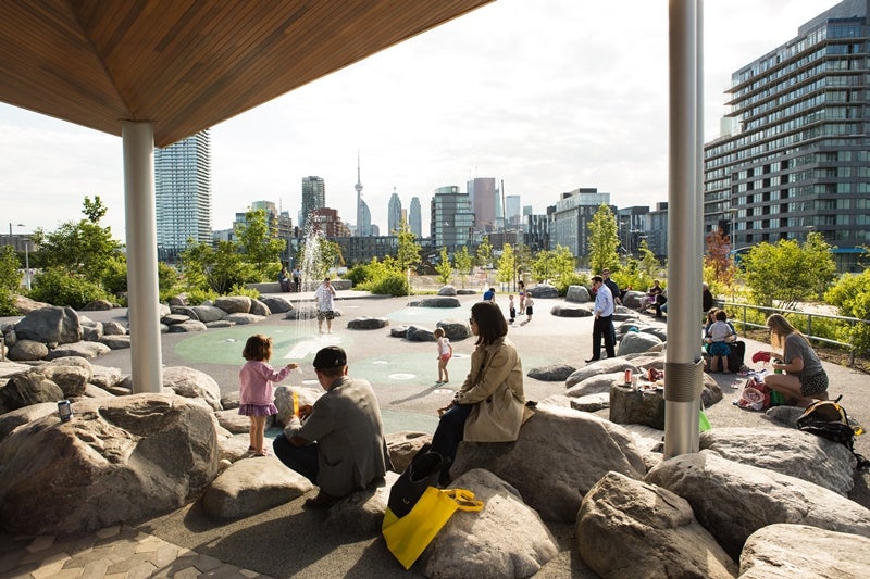 groups of people enjoying a splashpad and sitting on rocks in a park with downtown skyline in the background.
