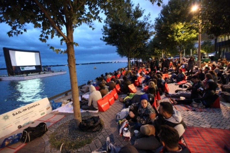 people sitting at the water's edge with a large movie screen on a water barge