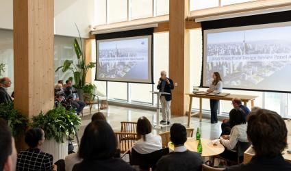 A person stands at a podium speaking to an audience in a modern conference room. Two large screens display a "2025 Waterfront Design Review Panel Awards" presentation, featuring an aerial cityscape image. Several people are seated, listening attentively.
