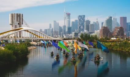  Photograph of a large group of canoes carrying colourful flags on tall poles, paddling down a waterway past a bridge with the Toronto skyline in the background.