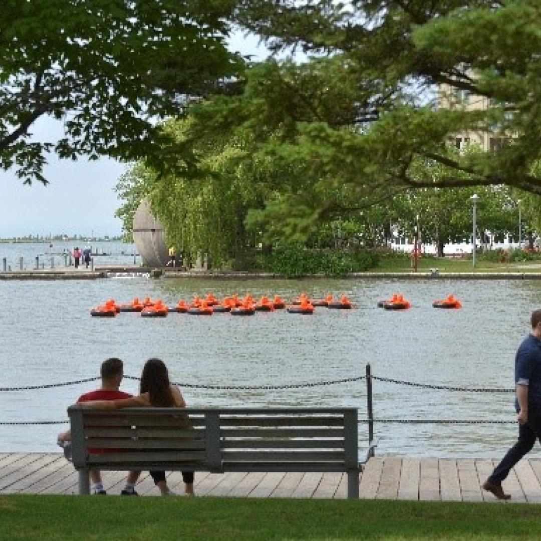 people sitting on a park bench at the water's edge looking out at Lake Ontario and a floating public art piece