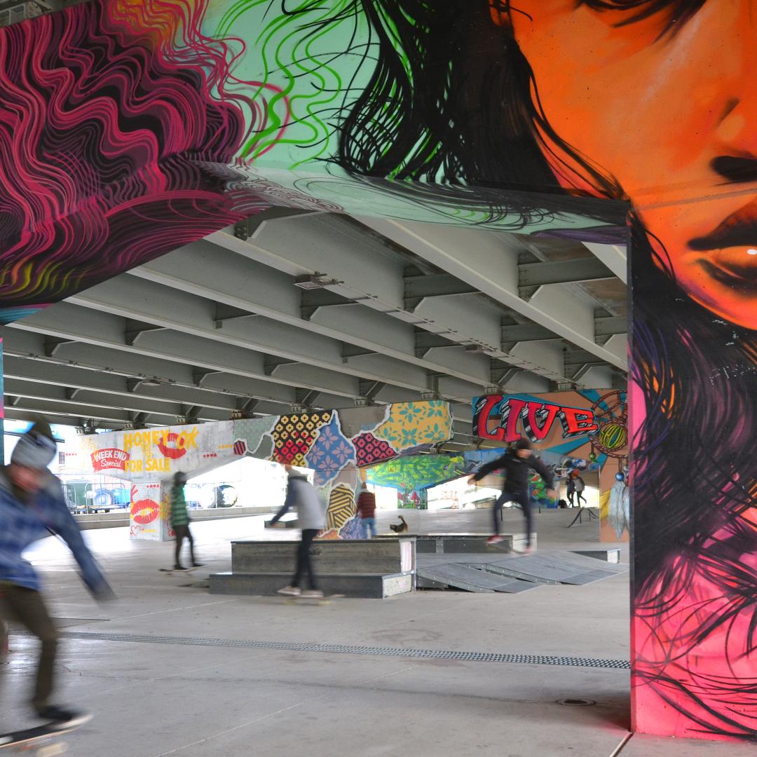 skateboarders enjoying an outdoor skate park underneath an overpass with beautiful public art murals on all the concrete pillars