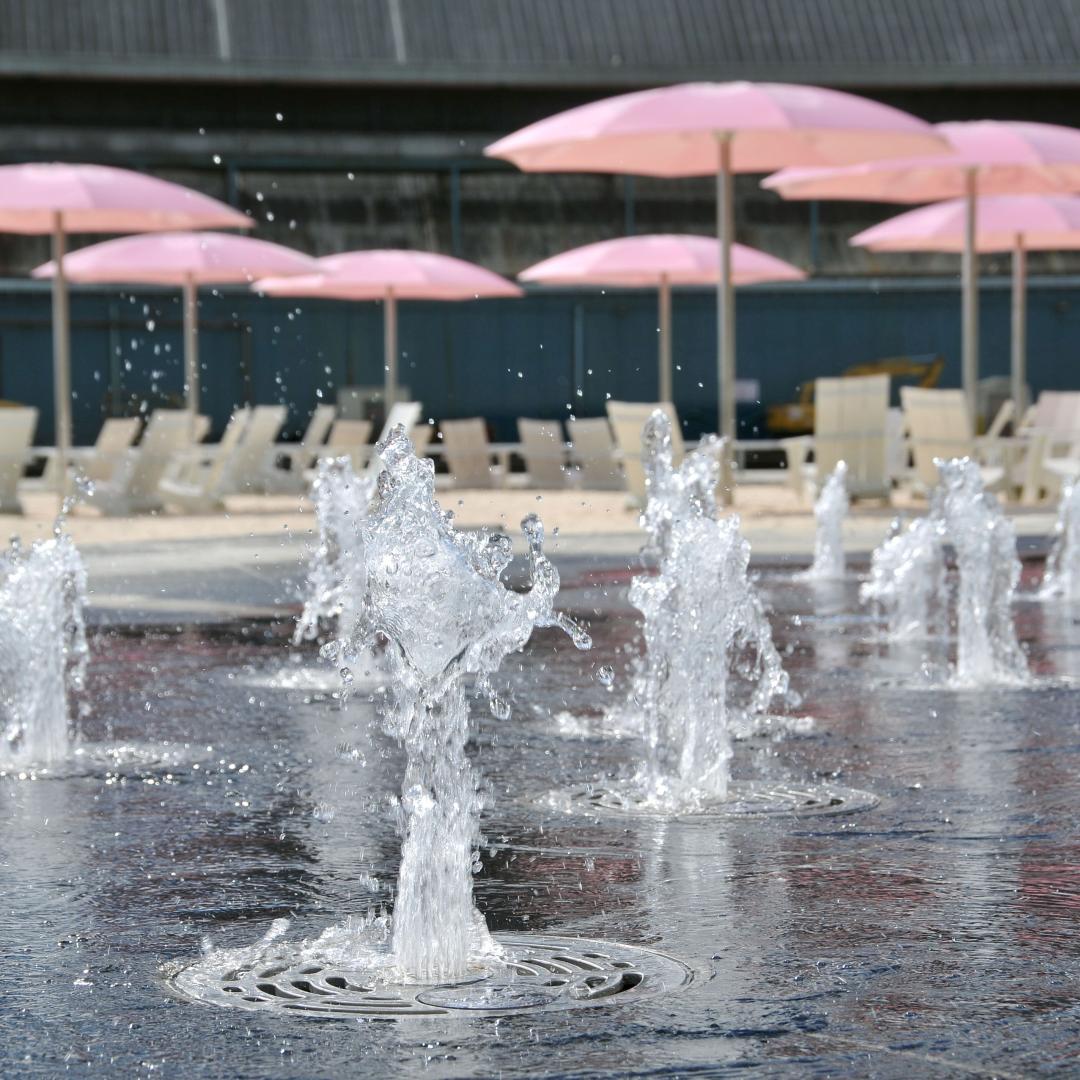 a close-up of water at a splash pad with pink umbrellas and sandy beach in the background